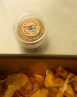 Close-up of crispy potato chips in a container with a round colorful sticker on the lid, placed on a surface.