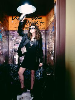 Person standing in a graffiti-covered room holding a can under a bright overhead light.