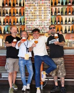 Four men standing at a bar holding glasses of craft beer with bottles displayed on shelves behind them at Open Baladin Roma.
