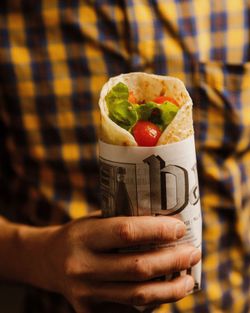 Panino avvolto in carta con pomodorini e lattuga, tenuto in mano da una persona con camicia a quadri gialla e blu.