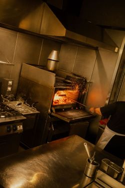 Open grill with flames inside a professional kitchen at Baladin Open Garden, showing part of a cook handling the fire.