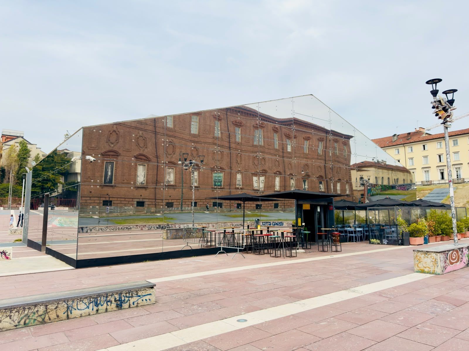 Façade miroir contemporaine réfléchissant un bâtiment historique, terrasse extérieure avec tables dans la place piétonne.