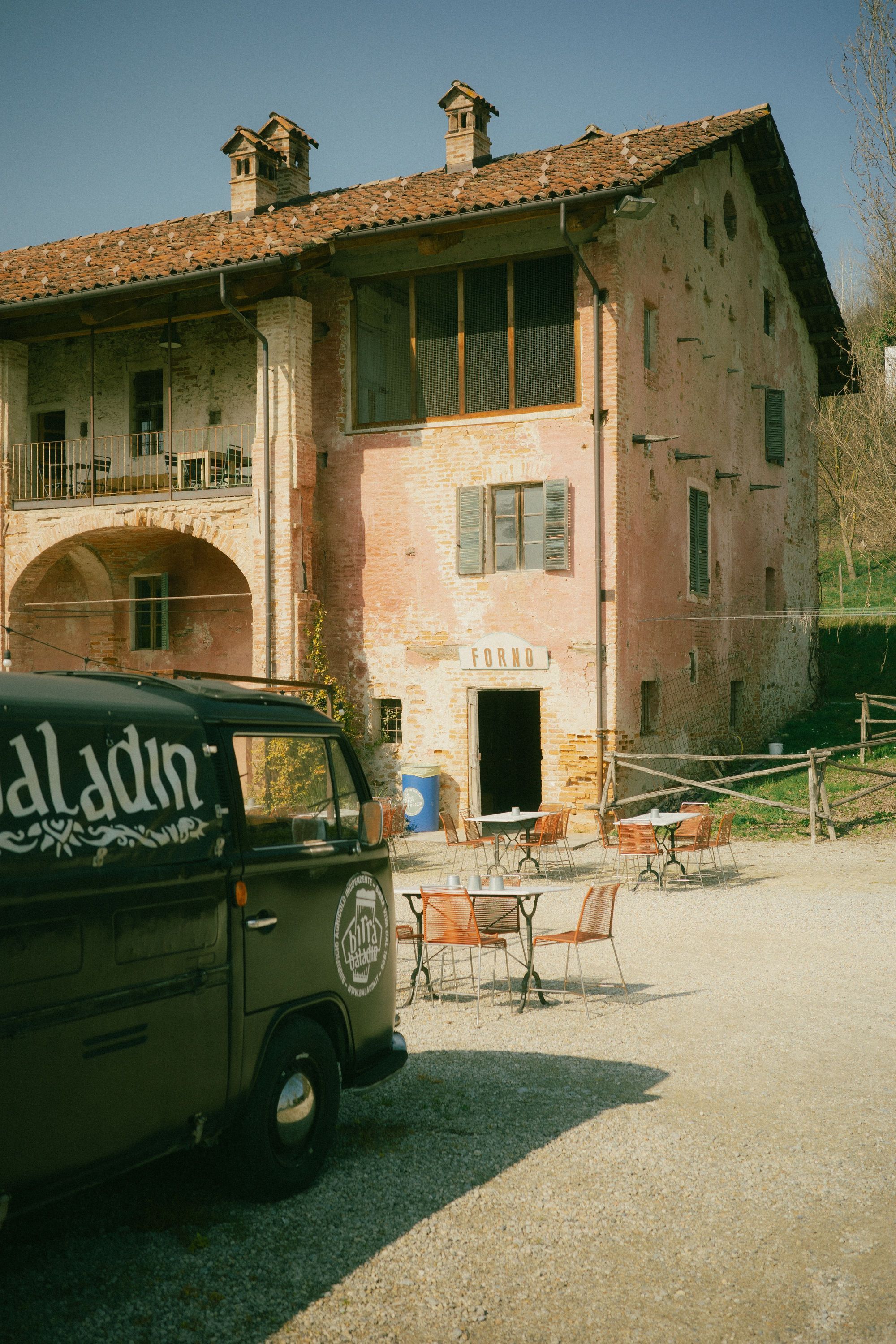 Façade d'une ancienne bâtisse en briques avec tables extérieures et un van Baladin stationné devant.