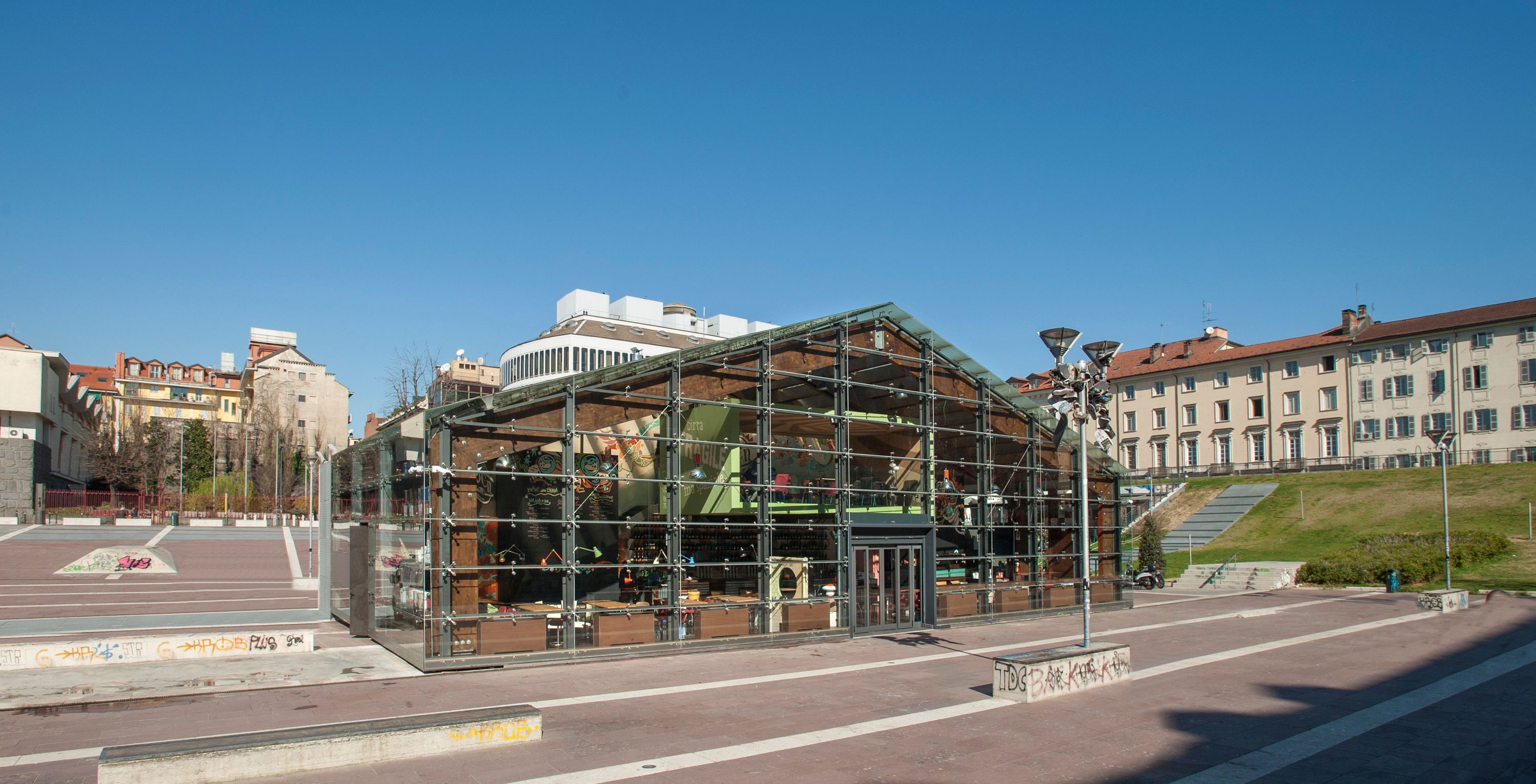 Modern glass building of Open Baladin Torino with outdoor seating area and urban surroundings on a clear day.