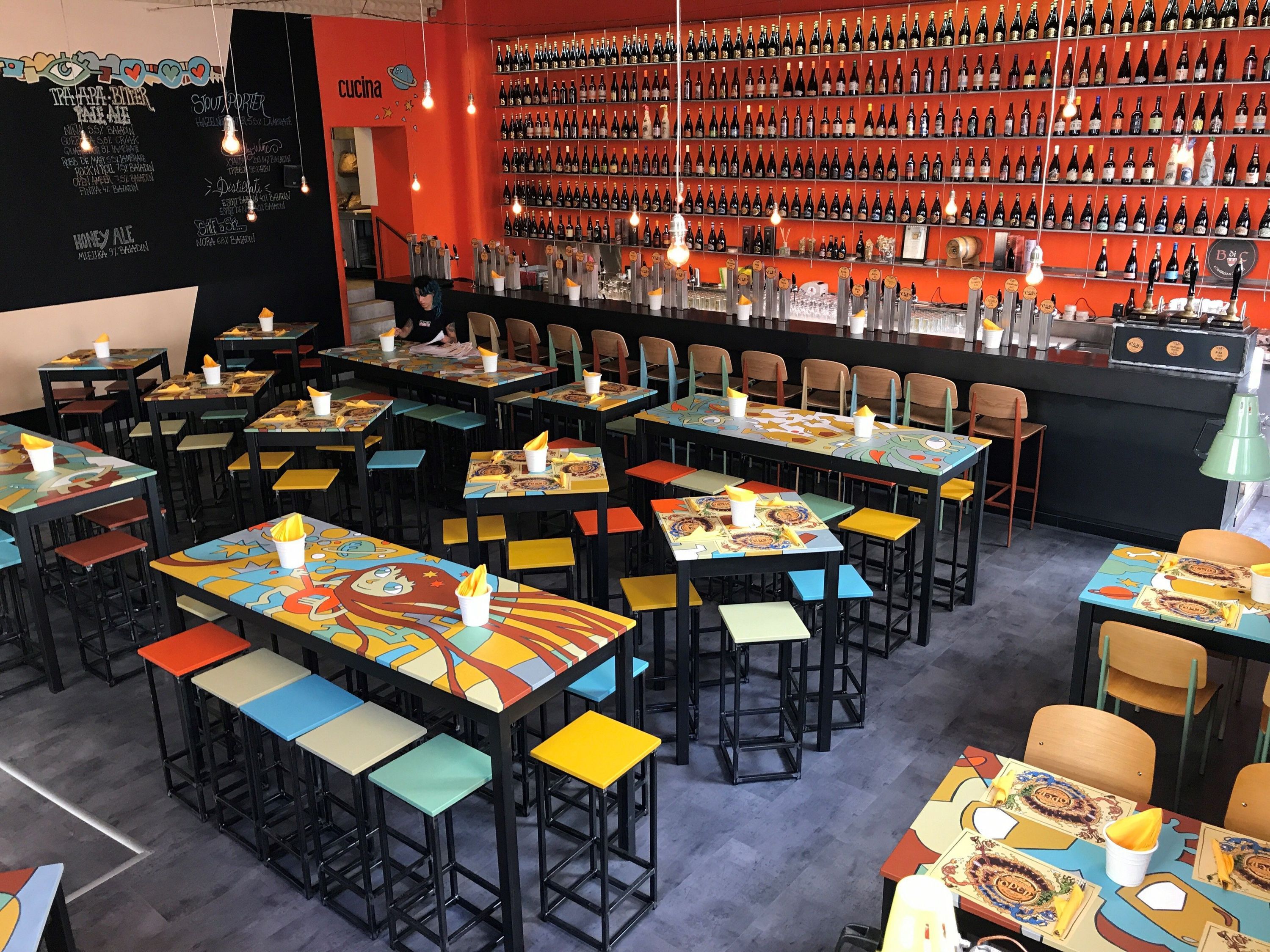 Interior of Open Baladin Roma featuring colorful tables, high stools, a long bar, and walls lined with craft beer bottles.