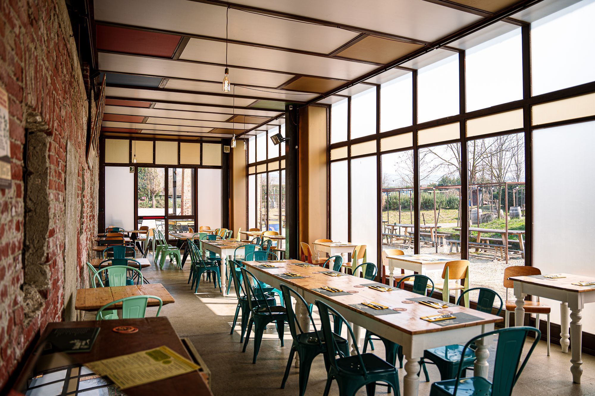 Spazio interno luminoso di Baladin Open Garden con tavoli e sedie verdi, vista sul giardino esterno.