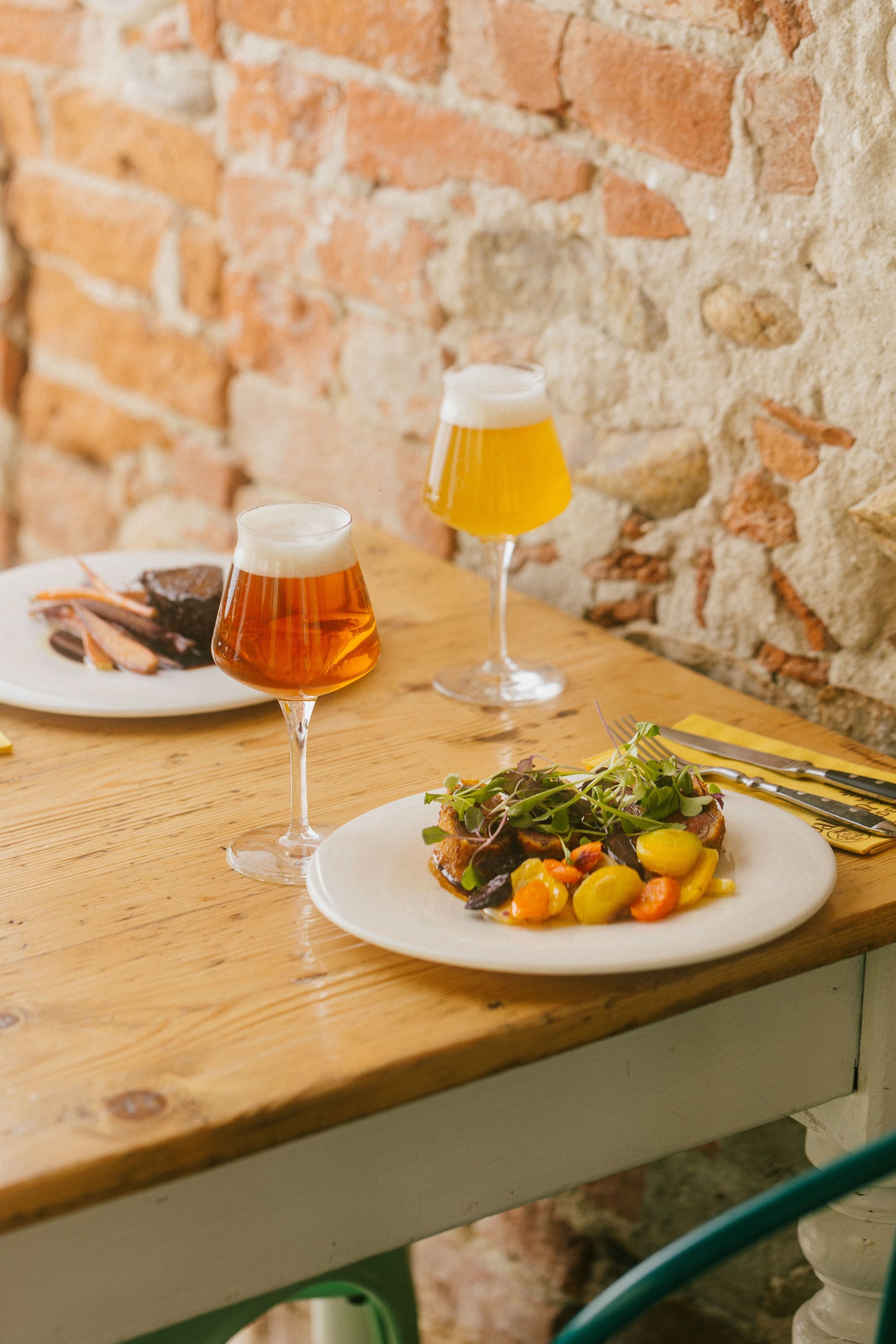 Zwei Gläser craft beer und zwei Teller mit Hamburger und Gemüse auf Holztisch vor Ziegelsteinmauer im Baladin Open Garden
