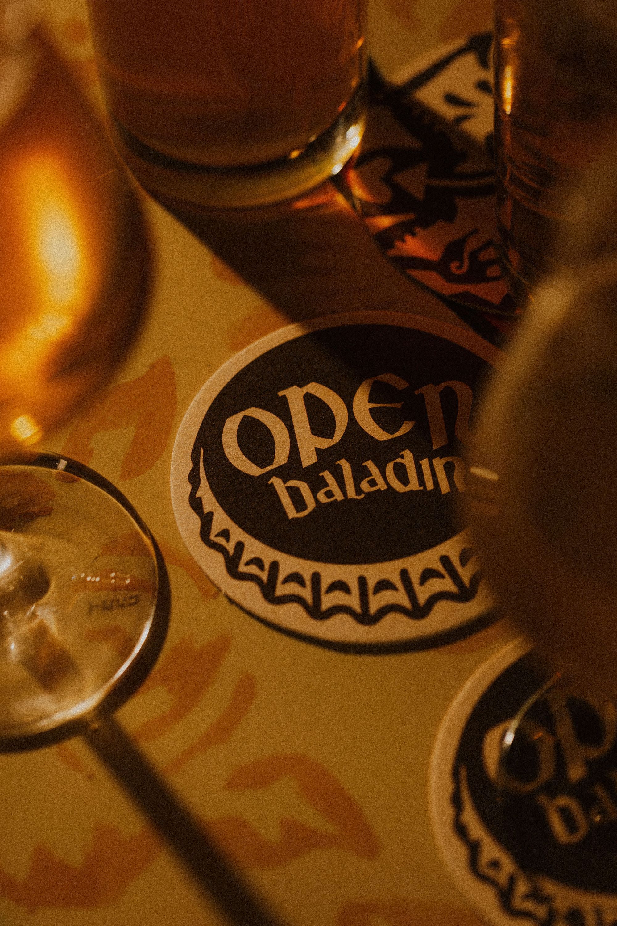 Tappetino per birra con logo Open Baladin circondato da bicchieri di birra artigianale su tavolo.