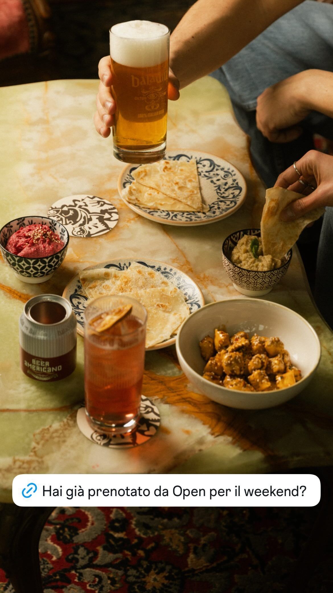 Verre de birra artigianale baladin servi avec des assiettes de snacks et un cocktail sur une table en marbre.