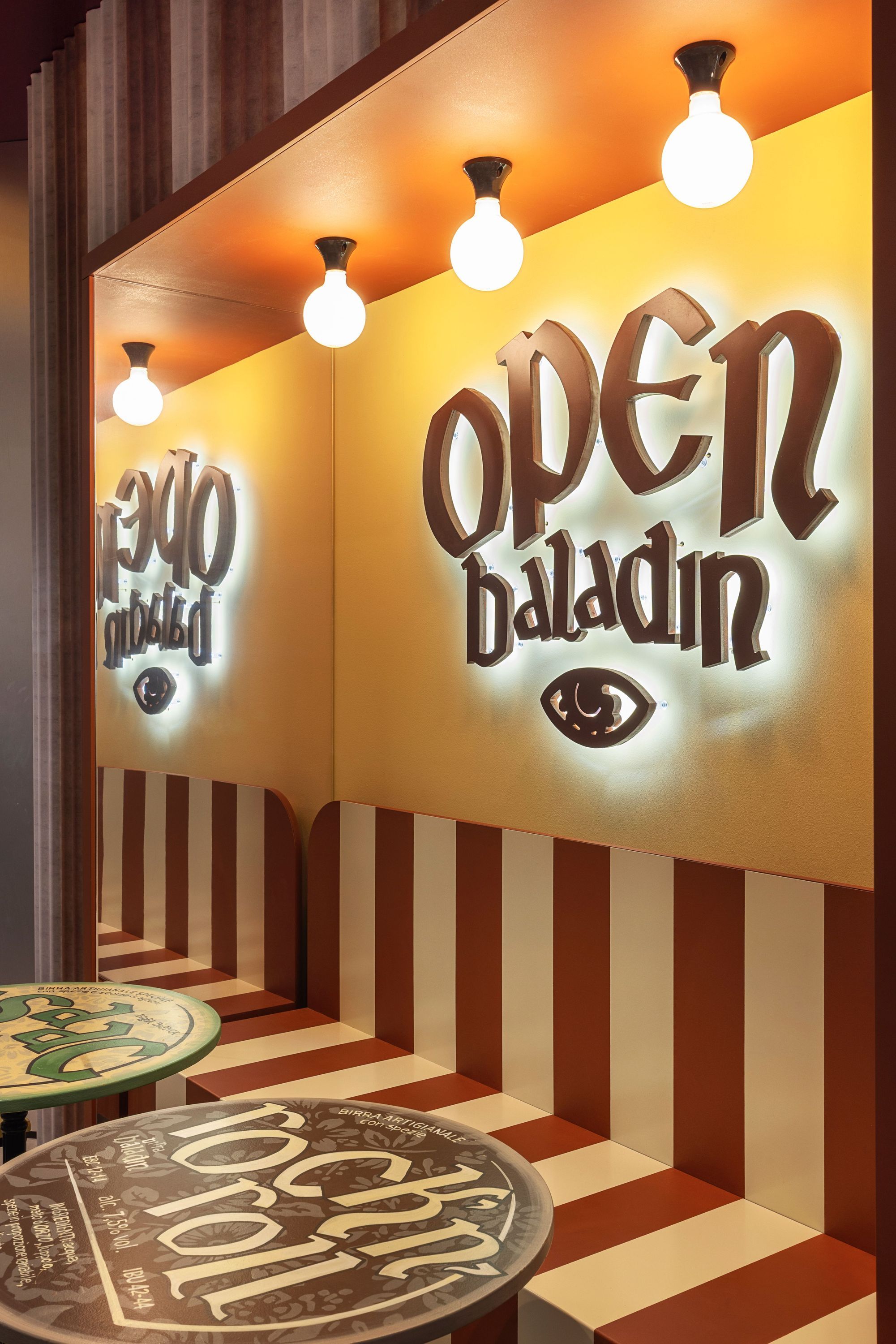 Logo illuminato di Open Baladin su parete con luci e mobili a strisce rosse e bianche nel locale.
