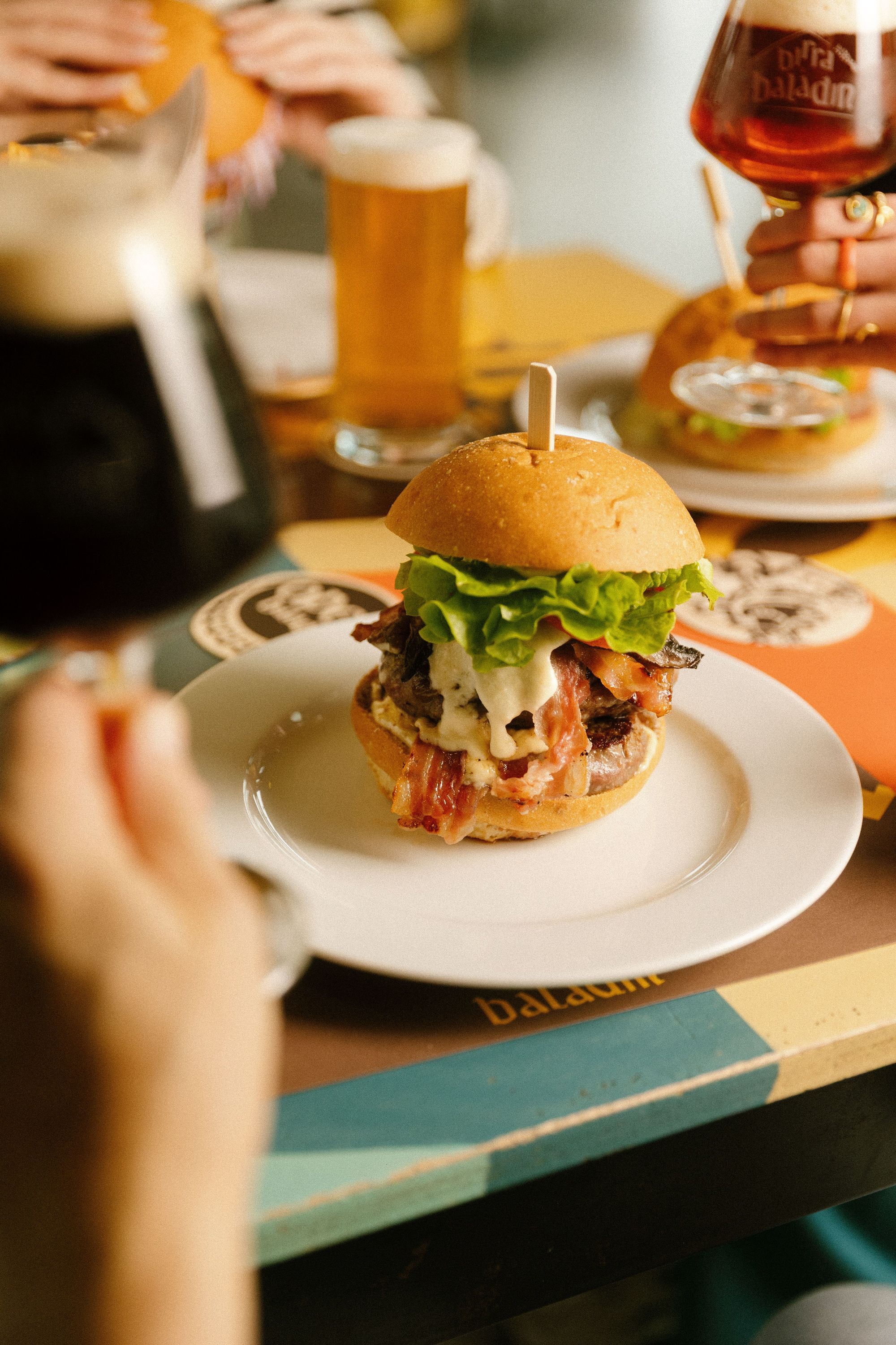 Gourmet hamburger topped with lettuce and melted cheese served on a white plate, accompanied by craft beer glasses.