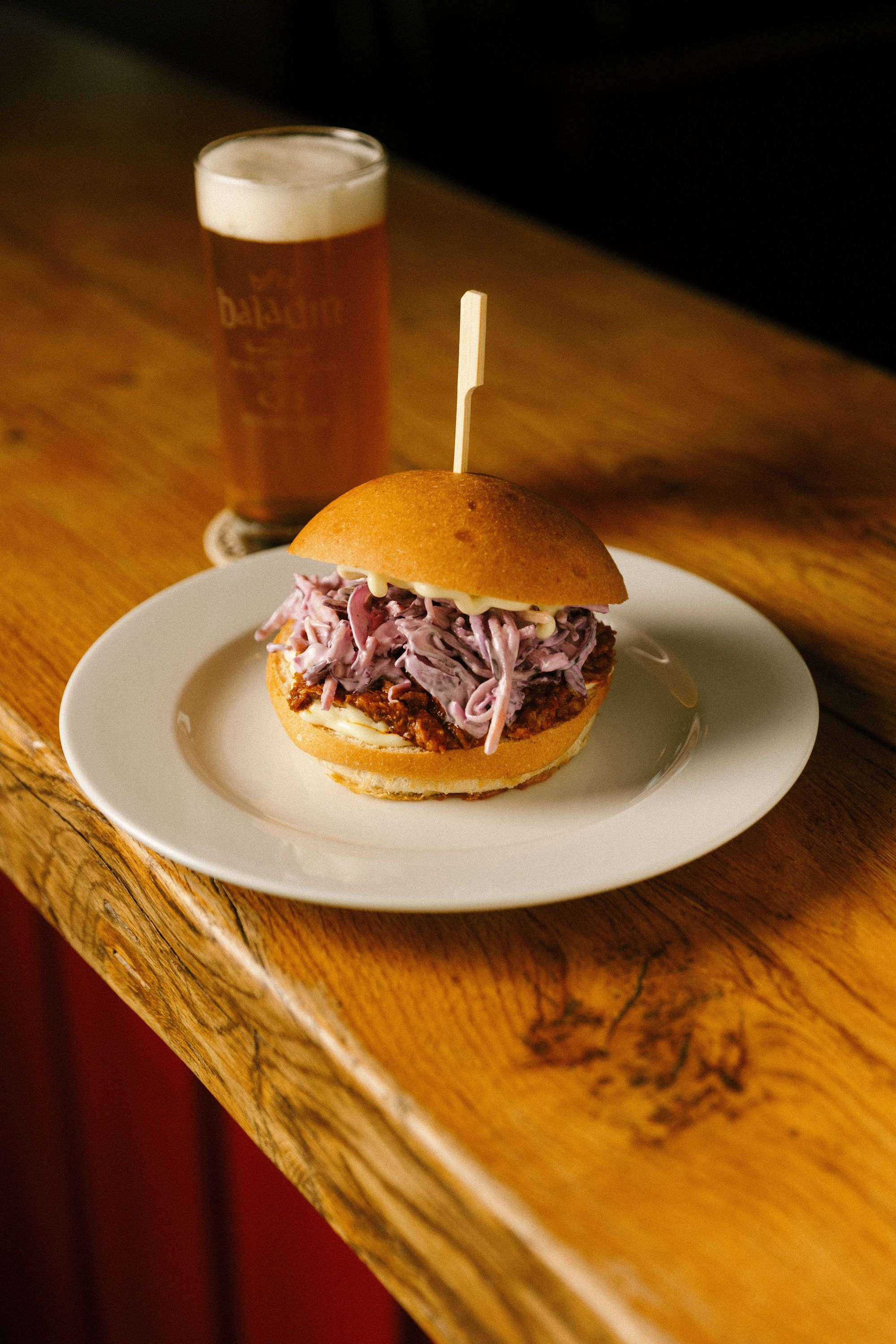 Hamburger topped with shredded purple cabbage on a white plate with a glass of Baladin craft beer on wooden surface.