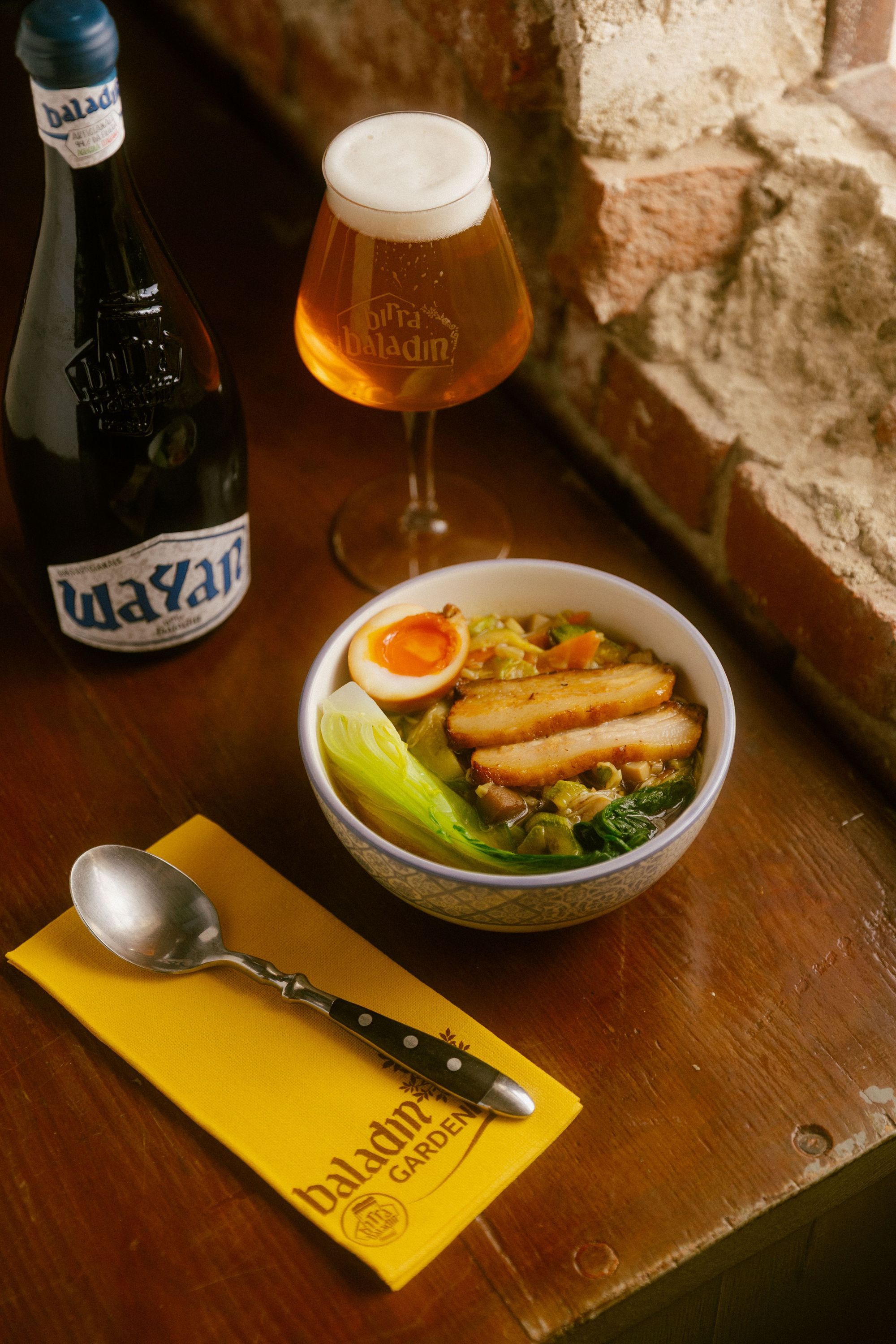 Ciotola di ramen con uovo, verdure e carne, accompagnata da birra artigianale Baladin su tavolo di legno rustico.