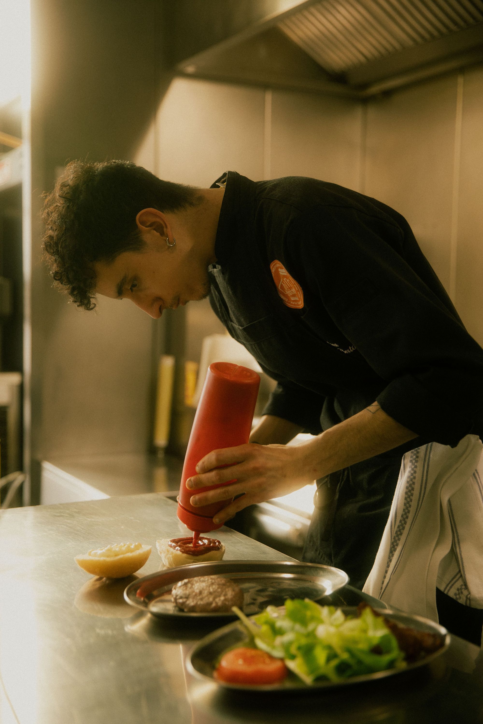Chef preparing a hamburger by squeezing ketchup onto the patty in a professional kitchen at Baladin Open Garden.