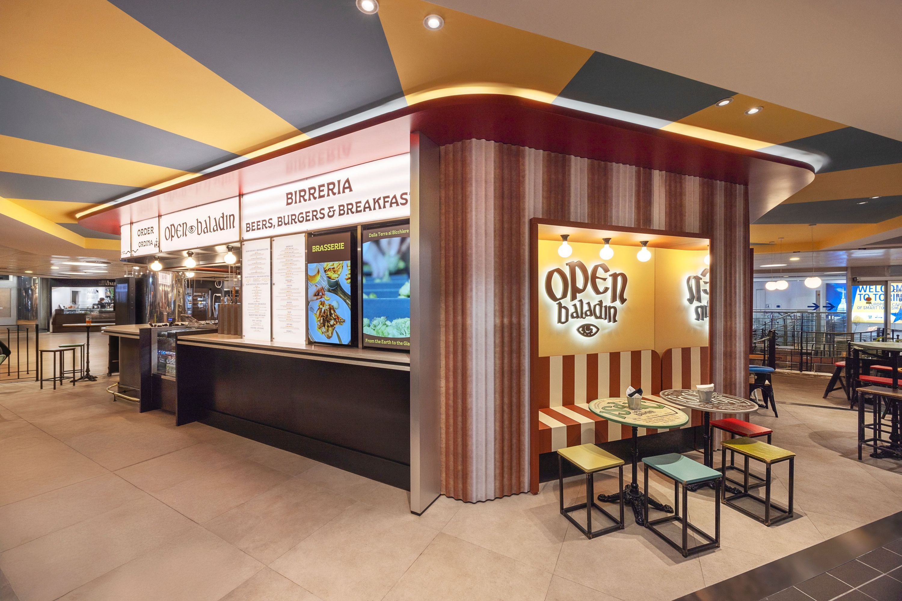 Locale Open Baladin Torino Aeroporto con area birreria, tavoli e sedie moderne, luce calda e design colorato al soffitto