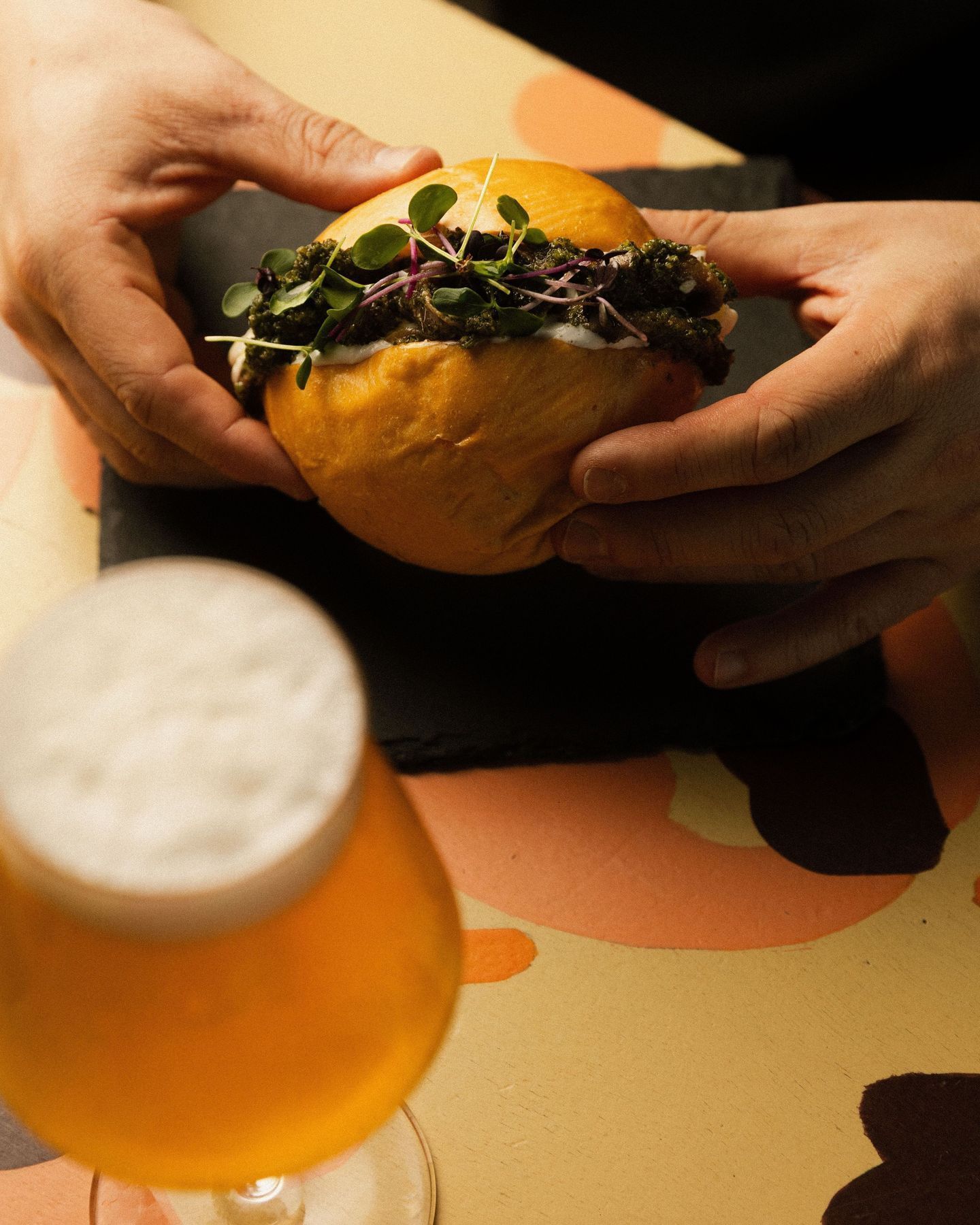 Hamburger mit frischen Kräutern, gehalten von zwei Händen, daneben ein Glas craft beer auf einem bunten Tisch.
