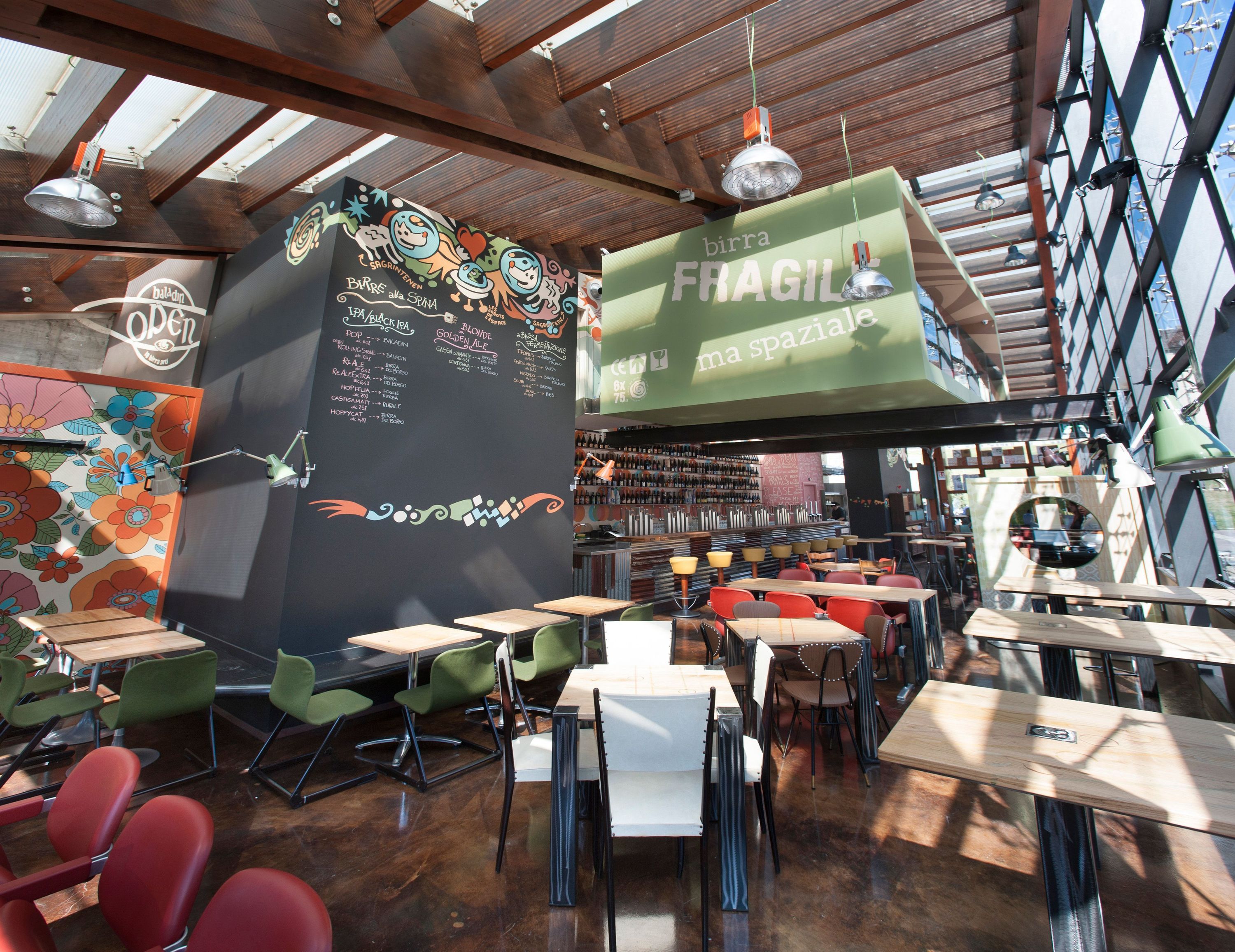 Intérieur lumineux d'Open Baladin Torino avec tables, chaises variées et un grand panneau vert pour la birra artigianale.