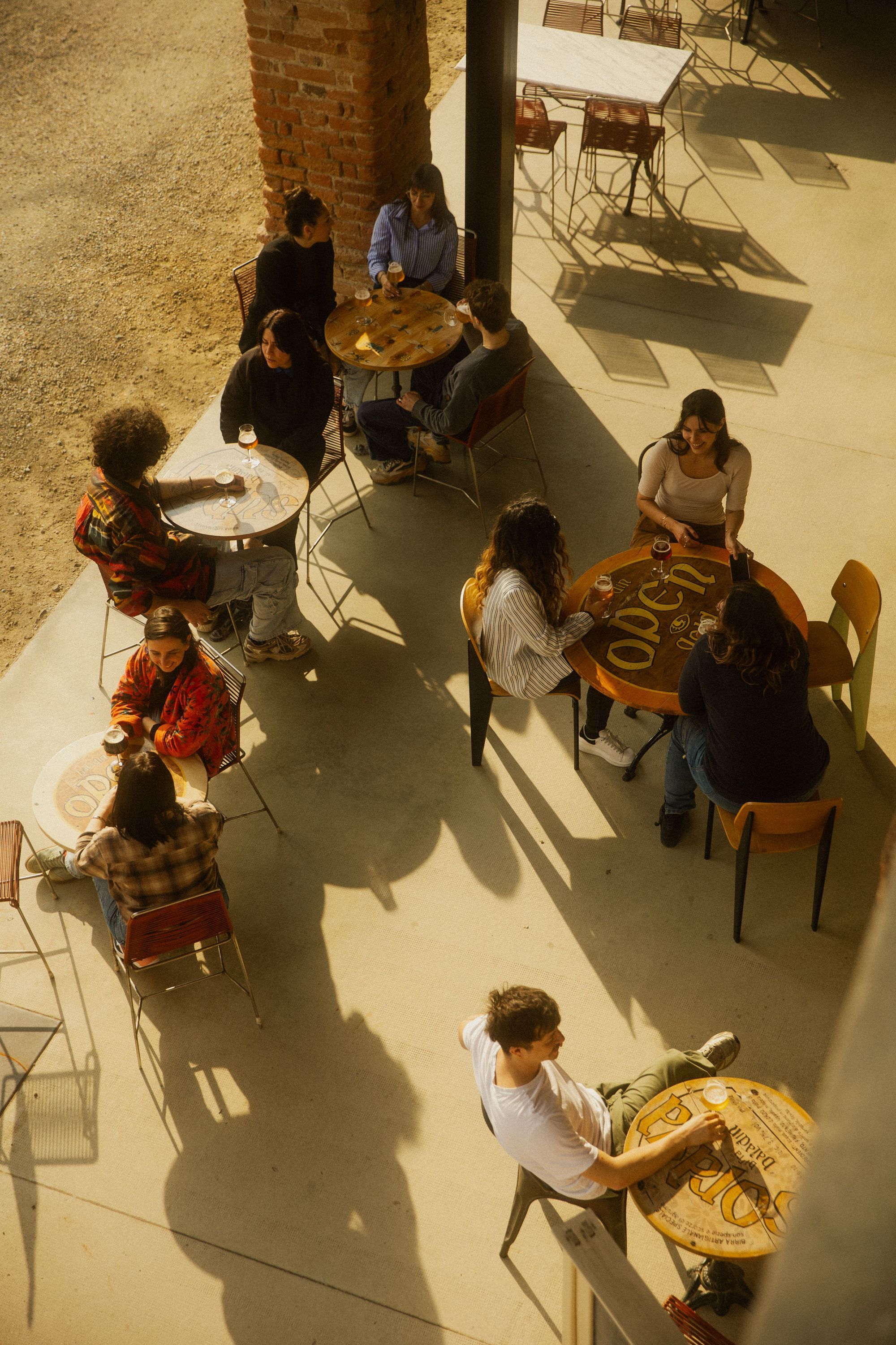 Patrons sitting at wooden tables under partial shade at Baladin Open Garden, enjoying drinks and socializing.