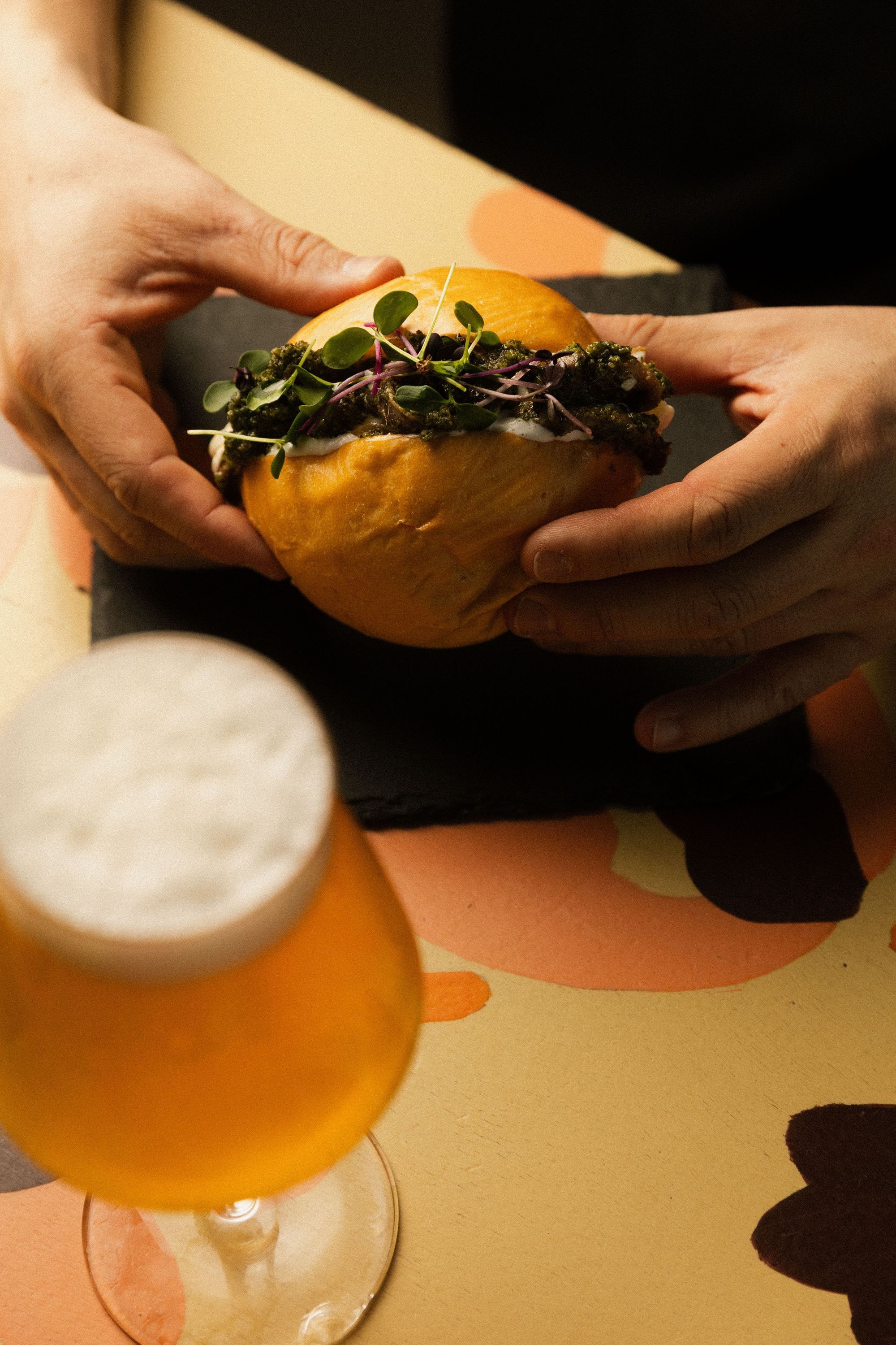 Hamburger gustoso con microgreens tenuto da mani, accanto a un bicchiere di birra artigianale chiara sul tavolo.