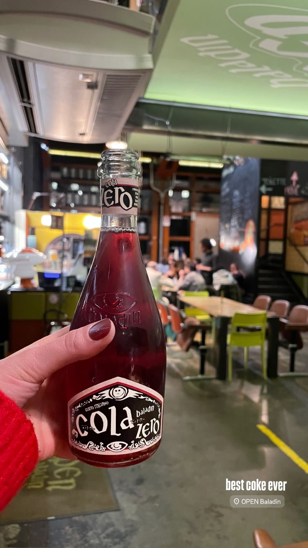 Craft beer bottle held inside Open Baladin Torino with a blurred background of tables and chairs in the bar area.