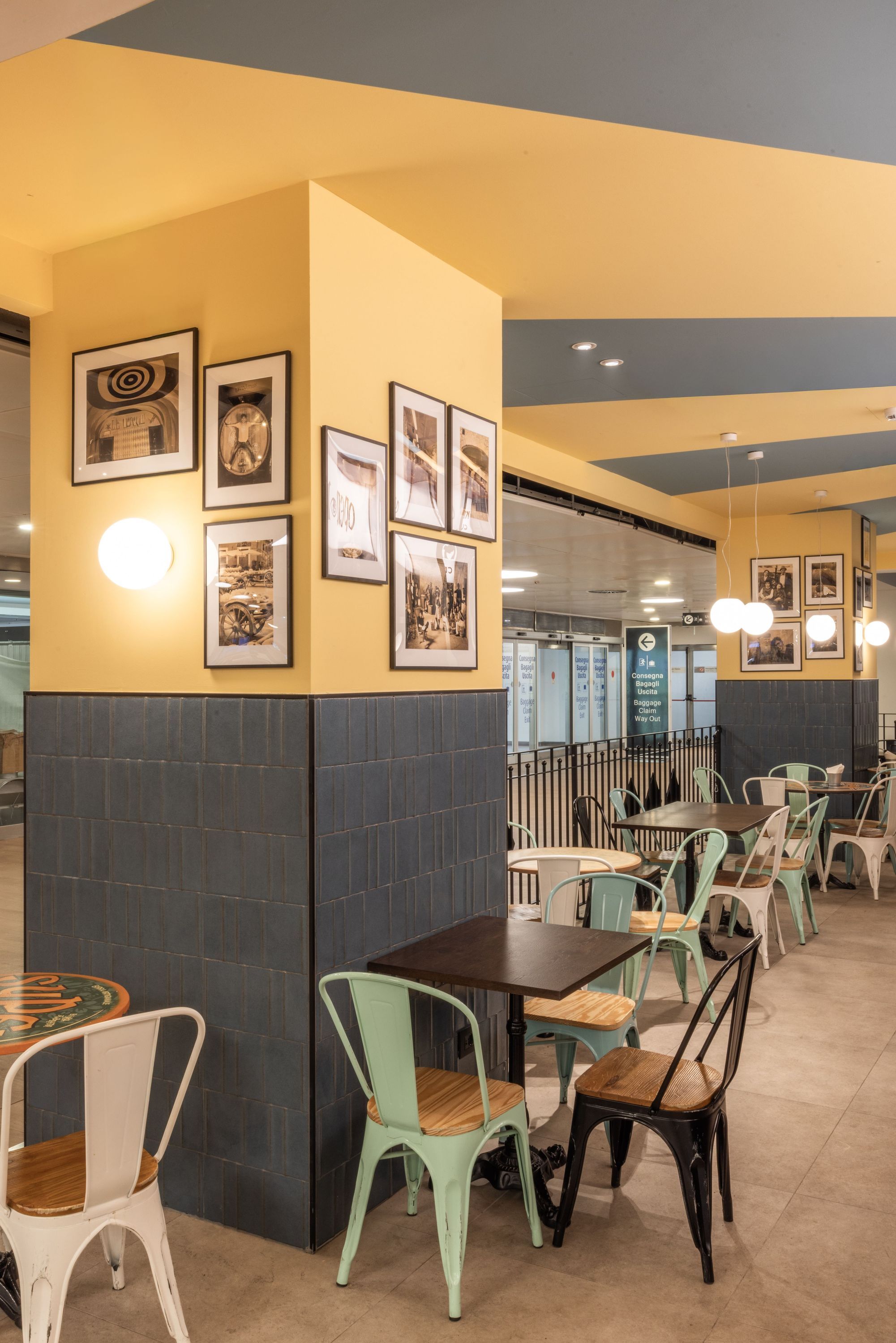 Interior of Open Baladin Torino Aeroporto with tables, chairs, yellow walls, and framed photos on columns.