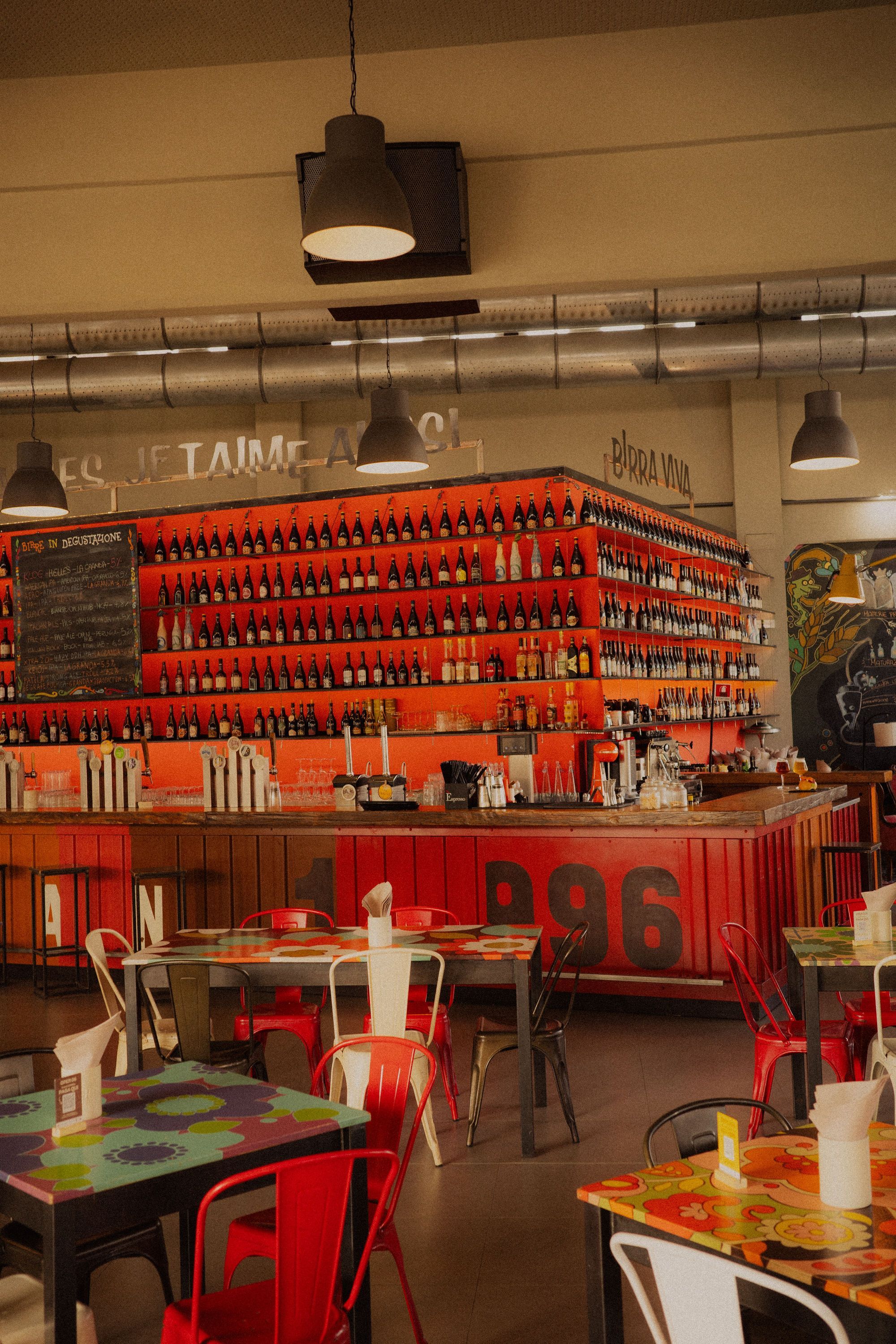 Bar area at Open Baladin Cuneo with shelves of craft beer bottles and red decor with tables and chairs.