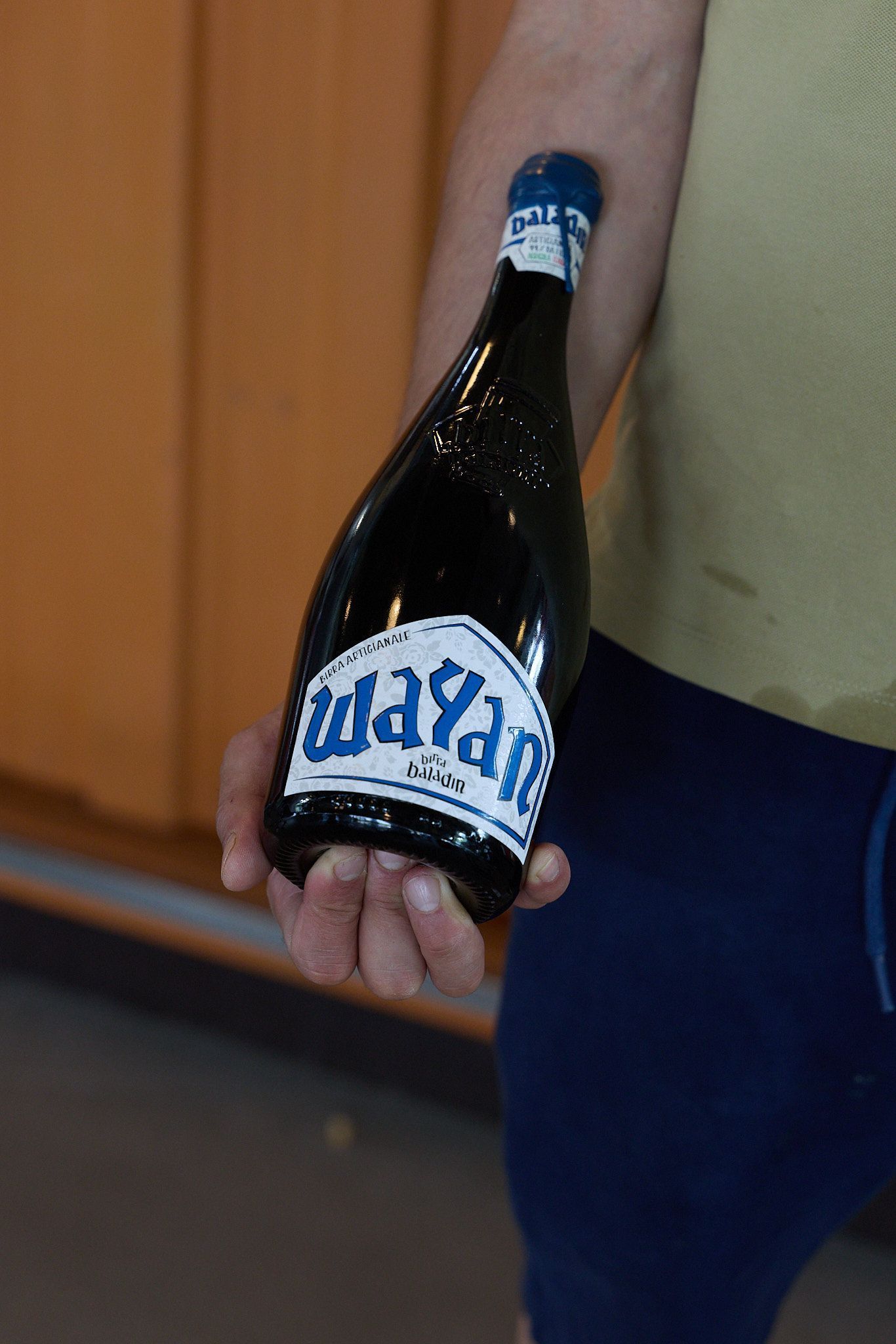 Person holding a dark bottle of Baladin craft beer labeled Wayan, with a blue and white label visible.